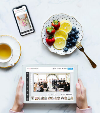 Hands editing a wedding photo album on a tablet while enjoying a plate of fruit and tea, with a phone displaying the same album.