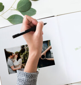 Hand writing in a wedding guestbook photo album, adding personal messages next to a couple’s photo.