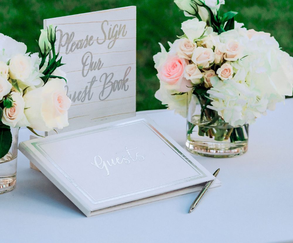 A wedding guest book with a sign reading "Please Sign Our Guest Book" surrounded by floral arrangements on a table, with a pen beside the book.