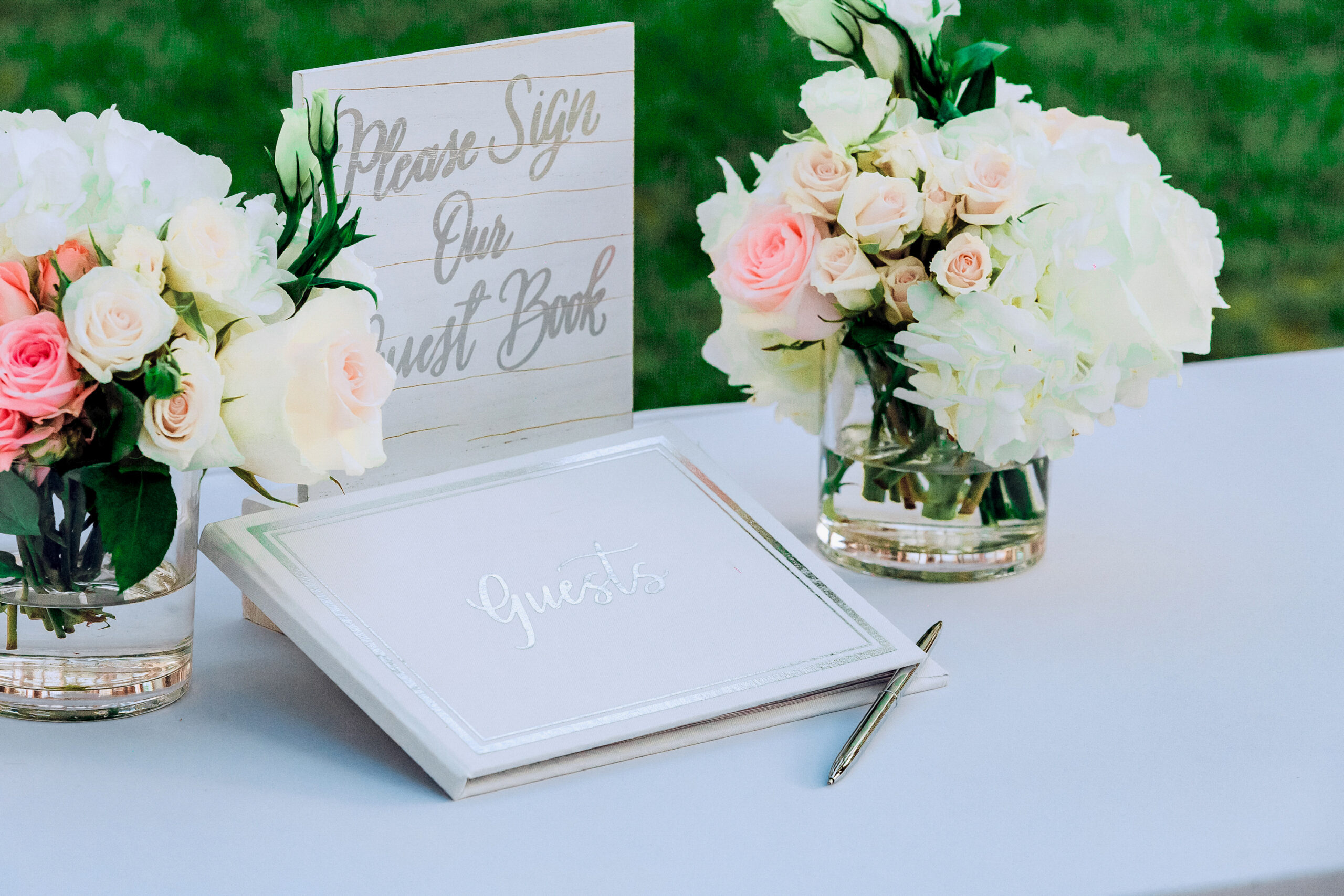 A wedding guest book with a sign reading "Please Sign Our Guest Book" surrounded by floral arrangements on a table, with a pen beside the book.