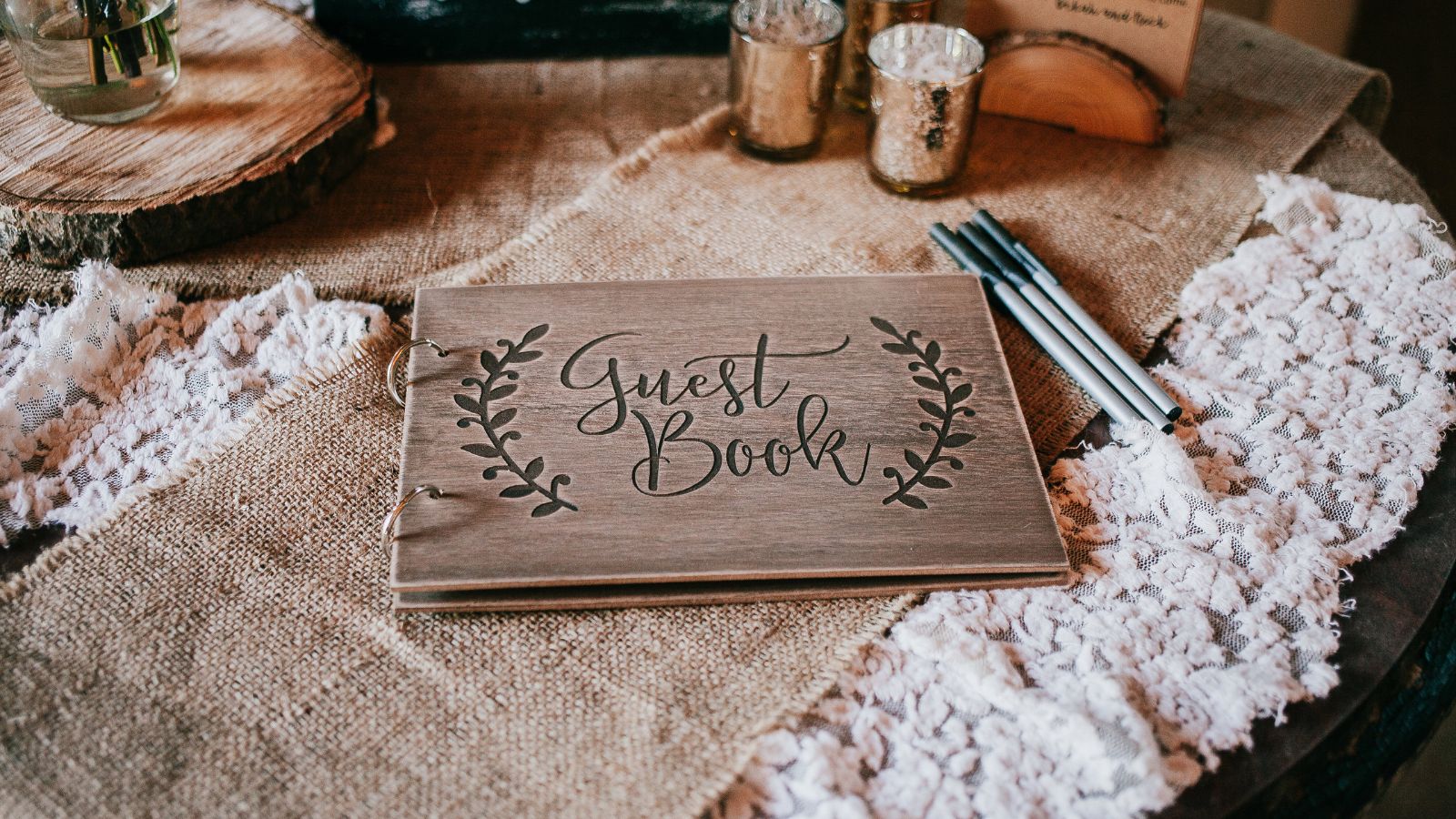 A wooden guest book, decorated with a floral design, sits on a table covered with burlap and lace. Pens are ready for guests to sign. The rustic, natural look suggests a wedding or similar event.