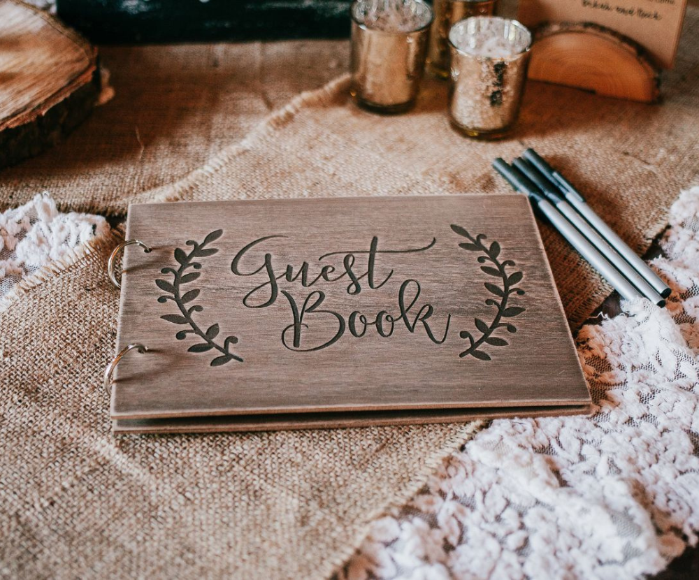 A wooden guest book, decorated with a floral design, sits on a table covered with burlap and lace. Pens are ready for guests to sign. The rustic, natural look suggests a wedding or similar event.