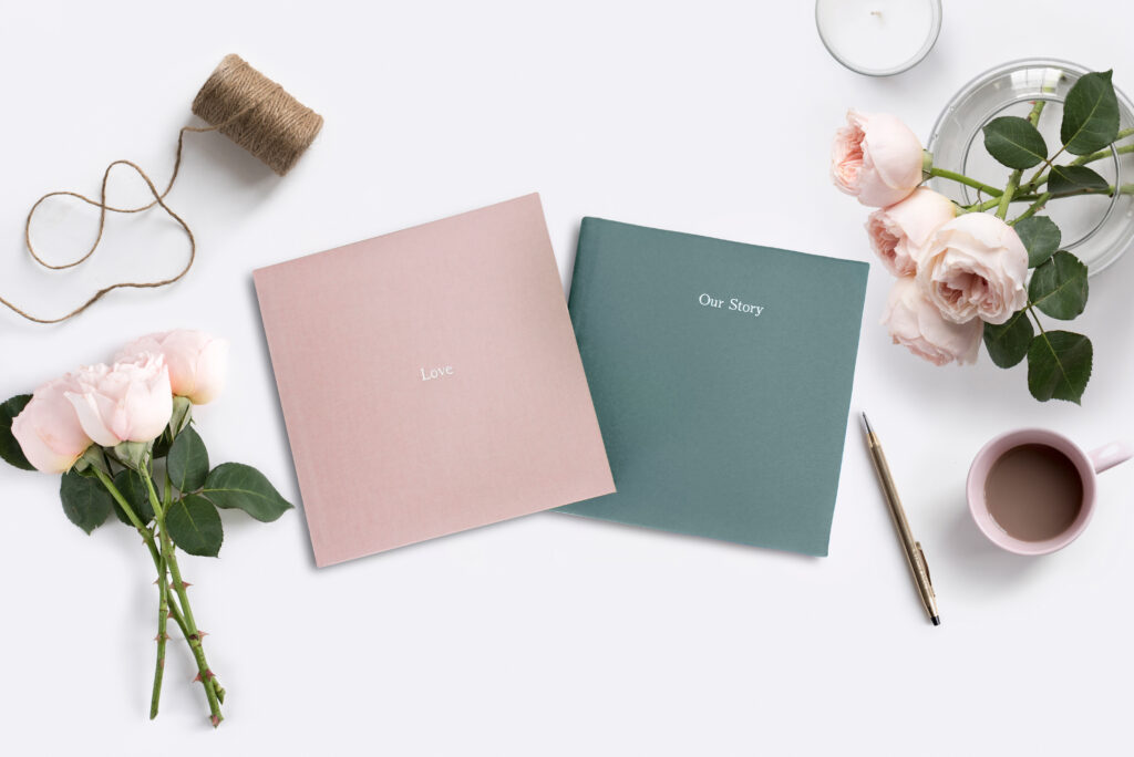 Two square photo albums, one pink with "Love" and one teal with "Our Story," are arranged on a white surface with roses, twine, a candle, and a pen.