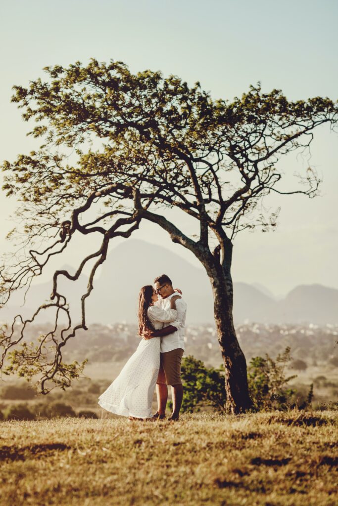 The couple is embracing under a large, sprawling tree in a scenic outdoor setting with golden grass and distant mountains in the background. The woman is wearing a flowing white dress, and the man is dressed in a casual shirt and shorts, suggesting a relaxed, natural vibe. The soft lighting and serene landscape add to the intimate, dreamy atmosphere of the photo.