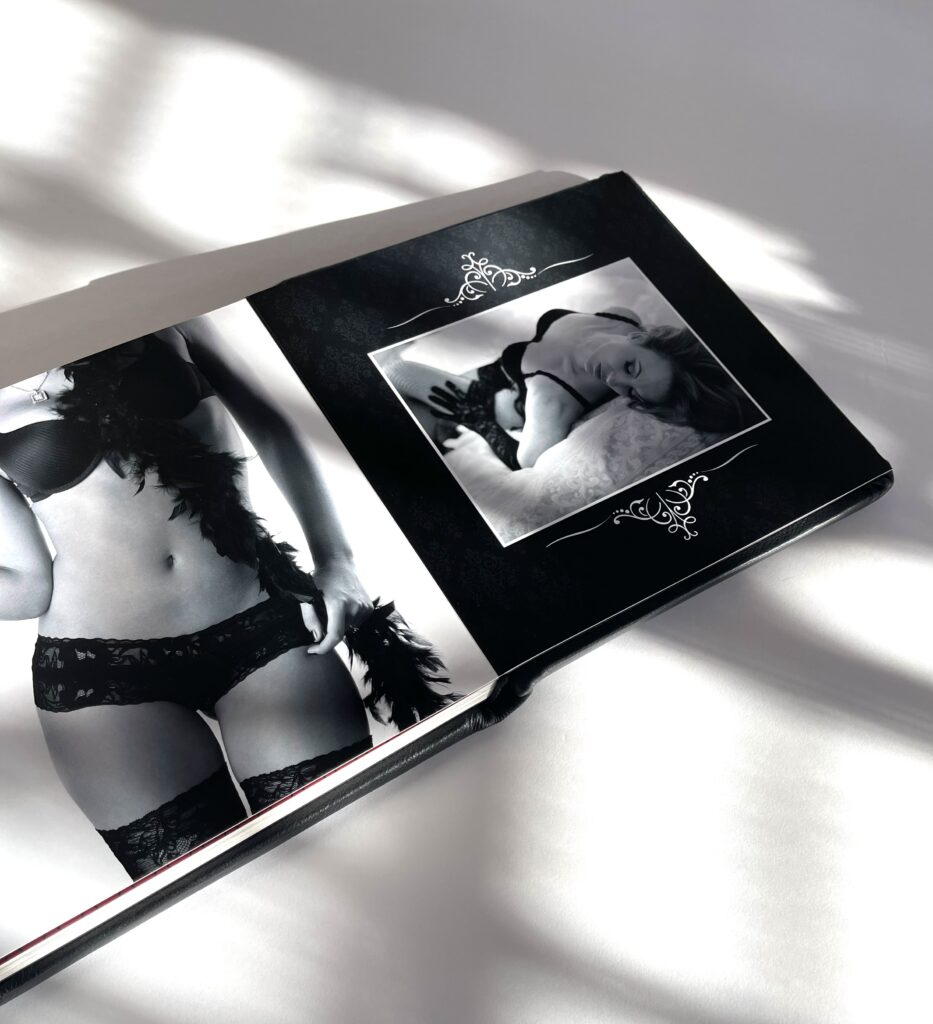 A boudoir book by My Bridal Pix lies open, showcasing a black-and-white photo of a woman in lace lingerie with feathers, her pose elegant. Shadows from sunlight add a soft touch to the intimate, stylish scene.