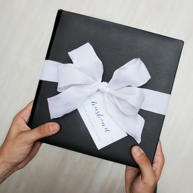 A person holding a black wedding album tied with a white bow and a "To Be Bound on Our Wedding Day" tag.