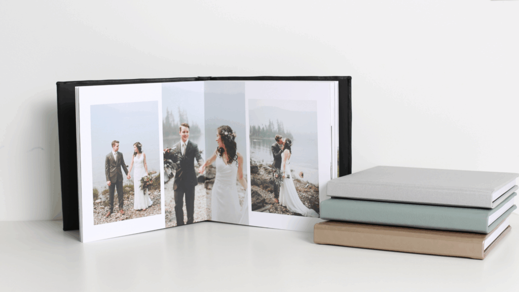 An open black wedding photo album with bride and groom images, next to a stack of closed albums in various colors.