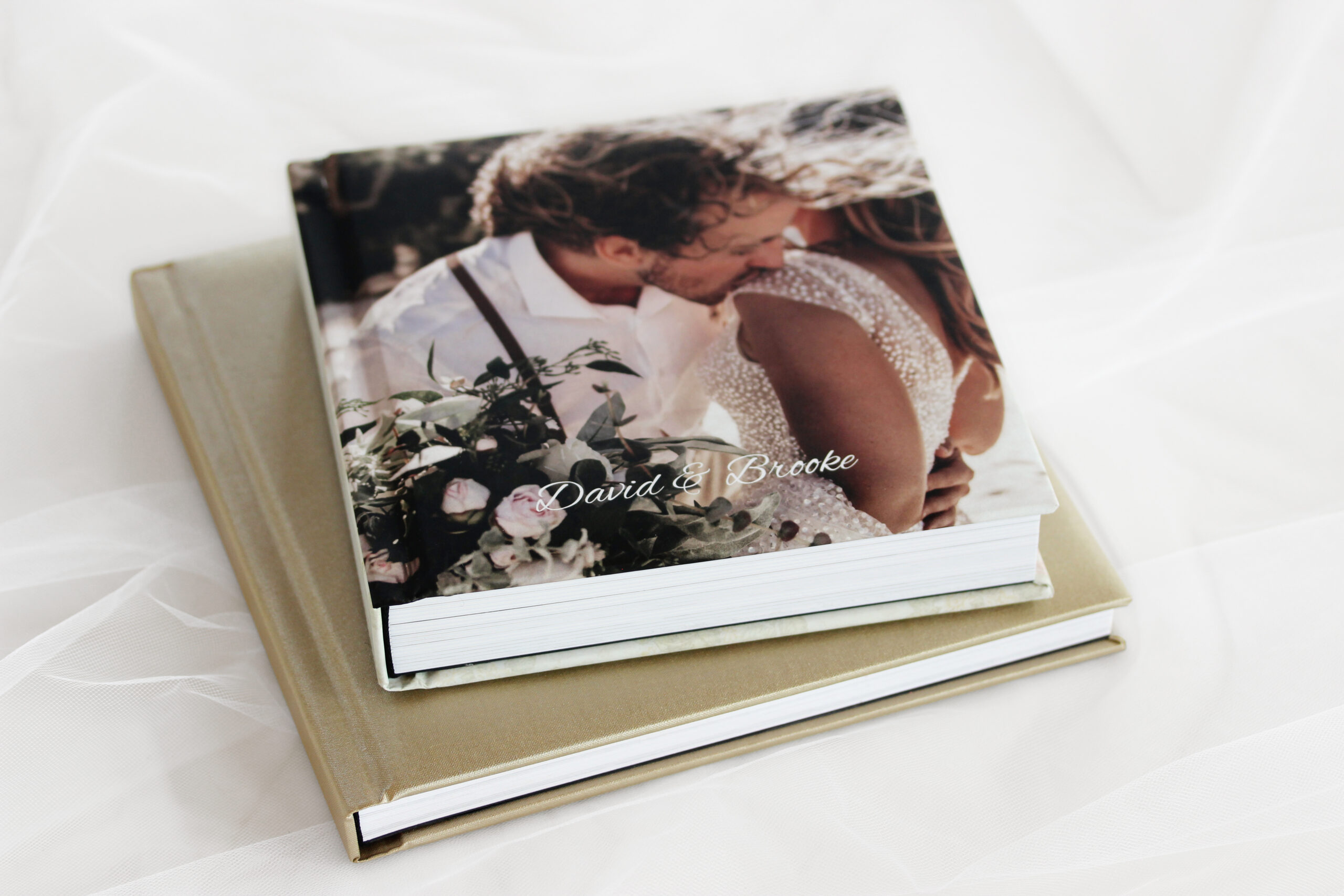 Wedding photo album with open pages showing bride and groom by a lake, surrounded by nature, with a stack of albums beside it.