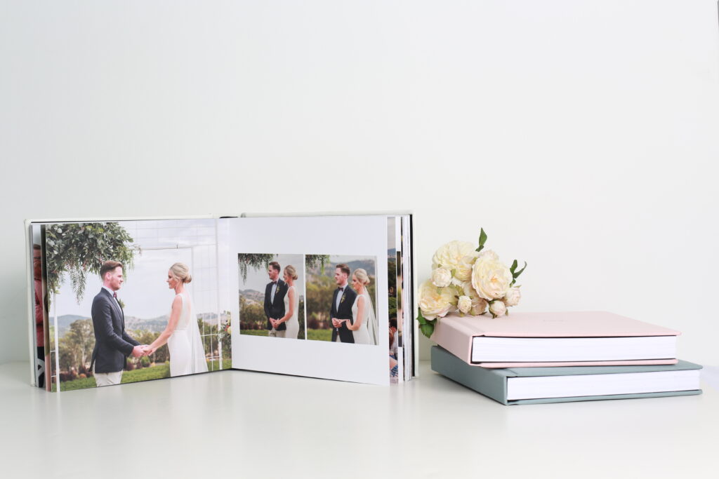 Wedding photo album open with bride and groom photos, next to stacked albums and flowers, capturing a romantic ceremony in a scenic setting.