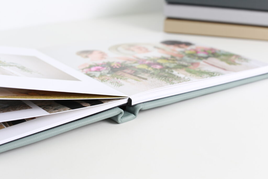 Wedding photo album open with group photos of bride, groom, and guests, next to a stack of albums on a white surface, capturing a joyful celebration.